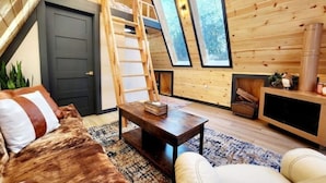 A cozy cabin interior with a loft bedroom, wooden walls, a large window, and a fireplace.