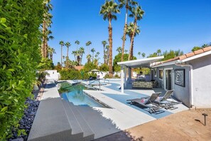 A pool area with lounge chairs, surrounded by palm trees and a building with a white facade. - Serenity Palms-Gorgeous Villa in Palm Springs (Palm Springs)