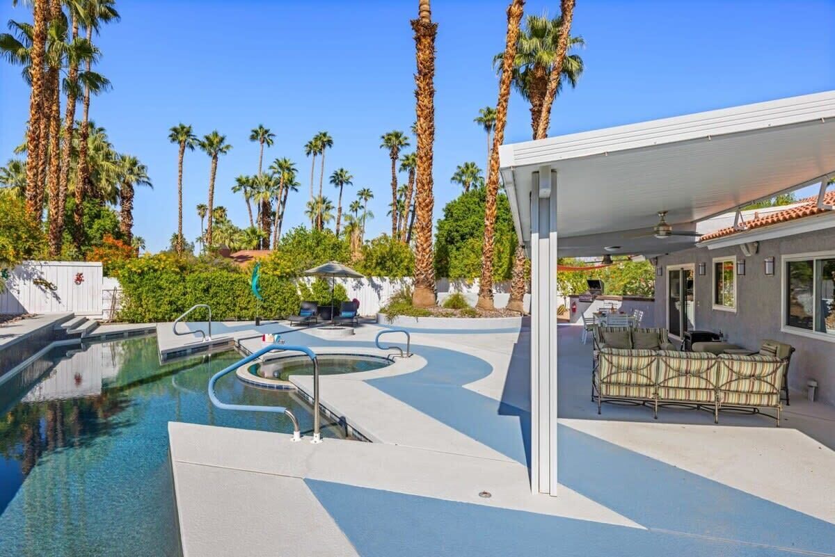 A swimming pool with a wavy edge, surrounded by palm trees and a covered seating area with a table and chairs.