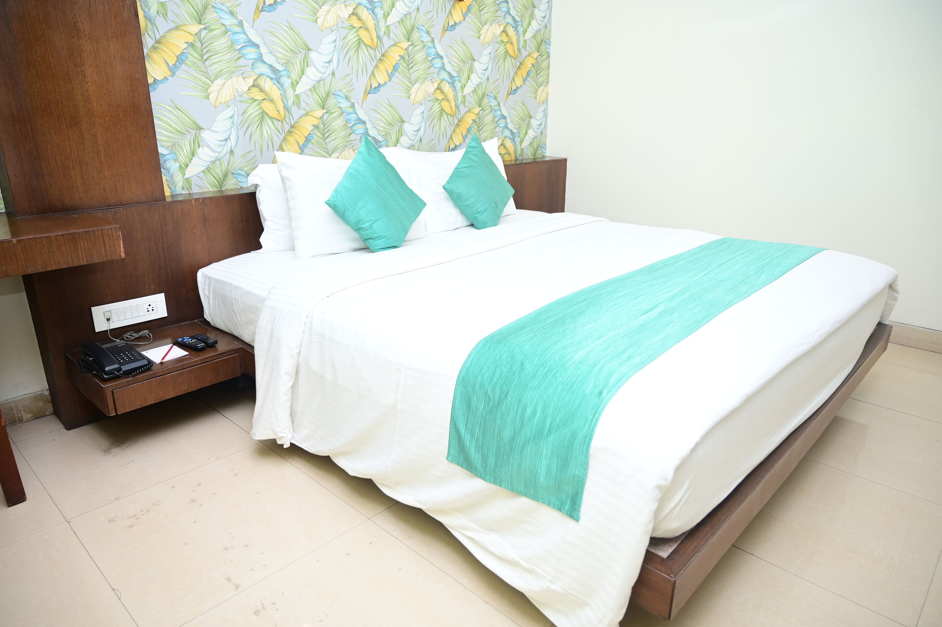 A neatly made bed with white bedding and teal accents, a wooden nightstand with a printer, and a wall with a tropical leaf pattern.