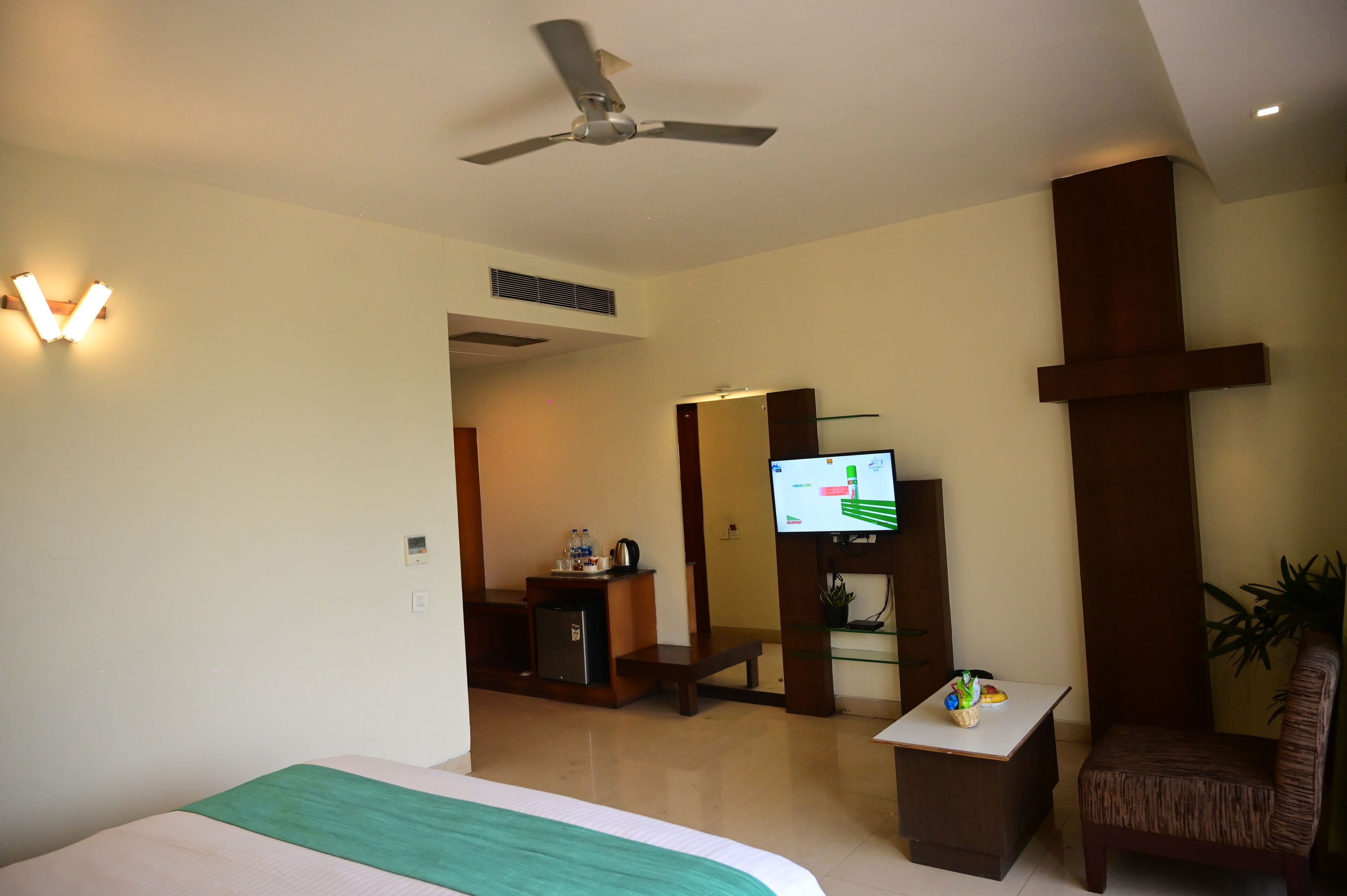 A hotel room with a bed, a desk, a chair, a television, a ceiling fan, and a cross on the wall.