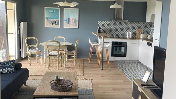 Una sala moderna con sofá, mesa de centro y comedor. Hay una cocina con salpicadero de azulejos y un televisor sobre un mueble de madera.