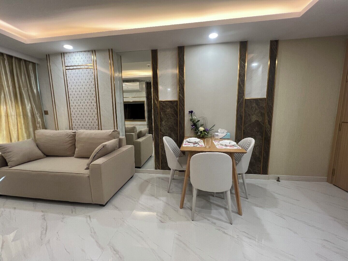 A modern living room with a beige sofa, a dining table set for four, and a TV in the background.