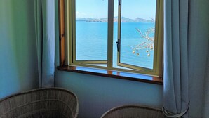 A room with a large window overlooking a body of water and a tree branch with white ornaments. There are two wicker chairs in the foreground. - Hôtel Hamaha Beach (Mamoudzou)