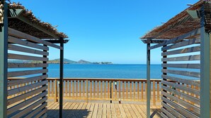 A wooden deck with a railing, leading to a beach with a clear view of the sea and a distant mountain. - Hôtel Hamaha Beach (Mamoudzou)
