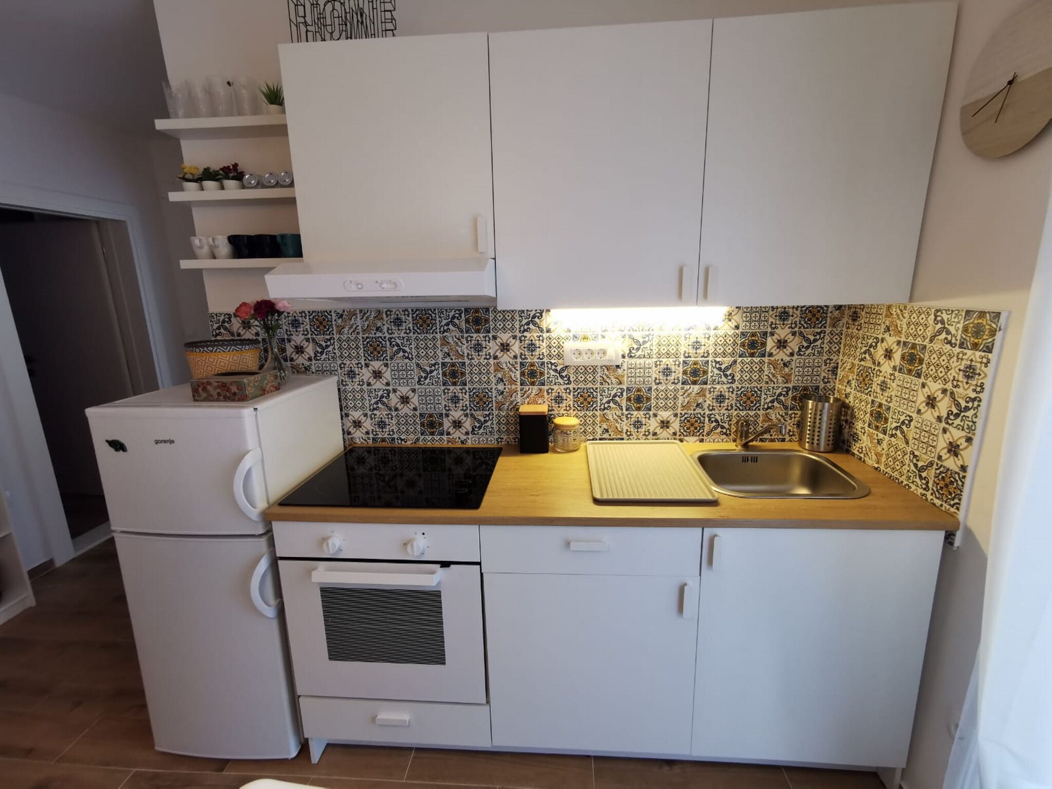 Kompakti keittiö, jossa on valkoiset kaapit, sisäänrakennettu uuni, valkoinen jääkaappi, pesuallas ja kuvioitu välitila.