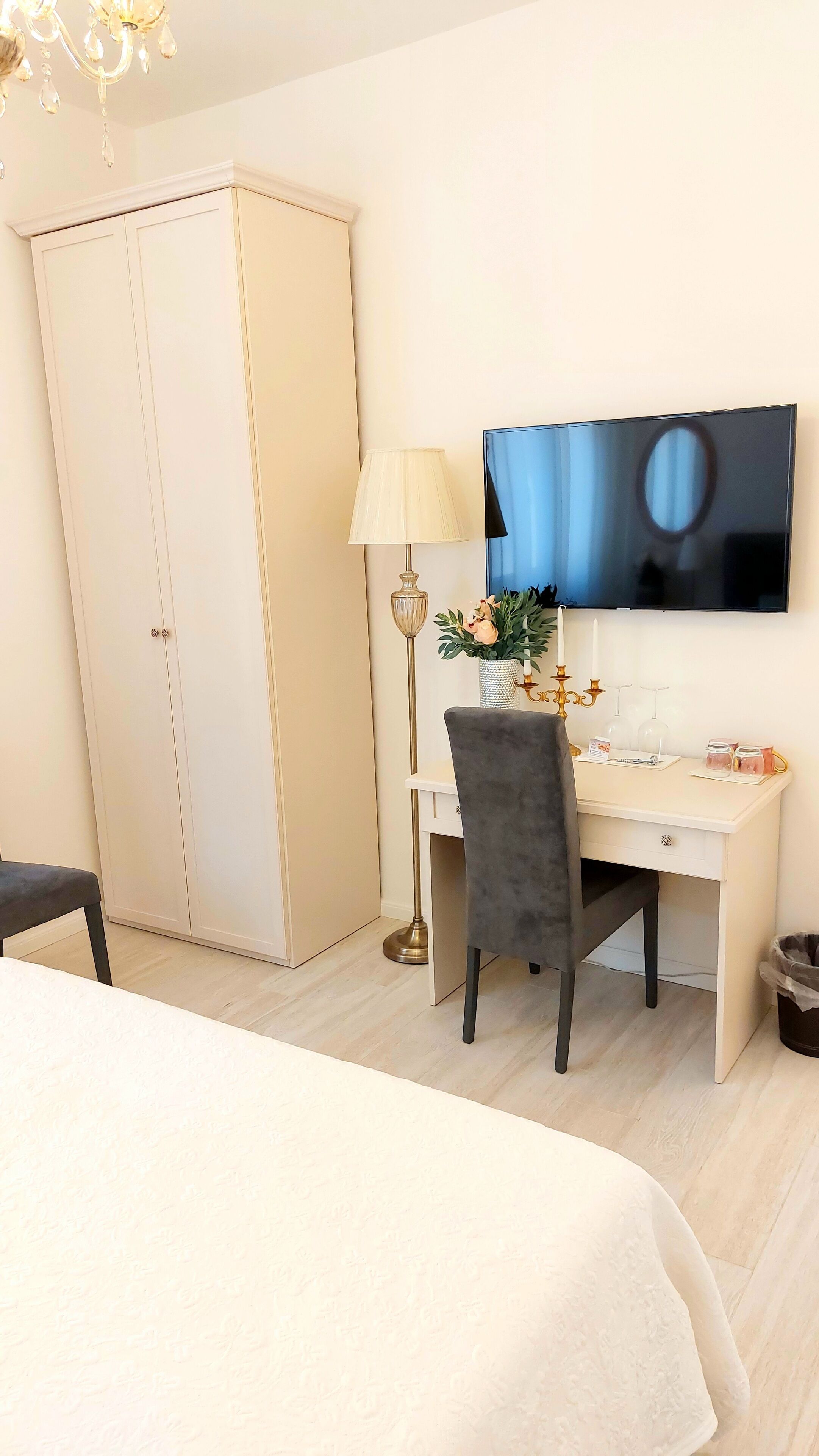 A hotel room with a bed, a desk with a chair, a TV, a wardrobe, and a floor lamp.