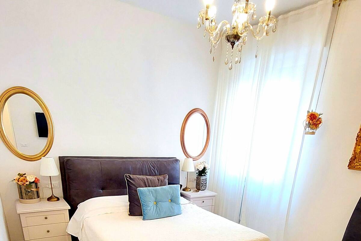 Quarto com cama grande, duas mesas de cabeceira, cômoda, cadeira, candelabro e dois espelhos na parede.