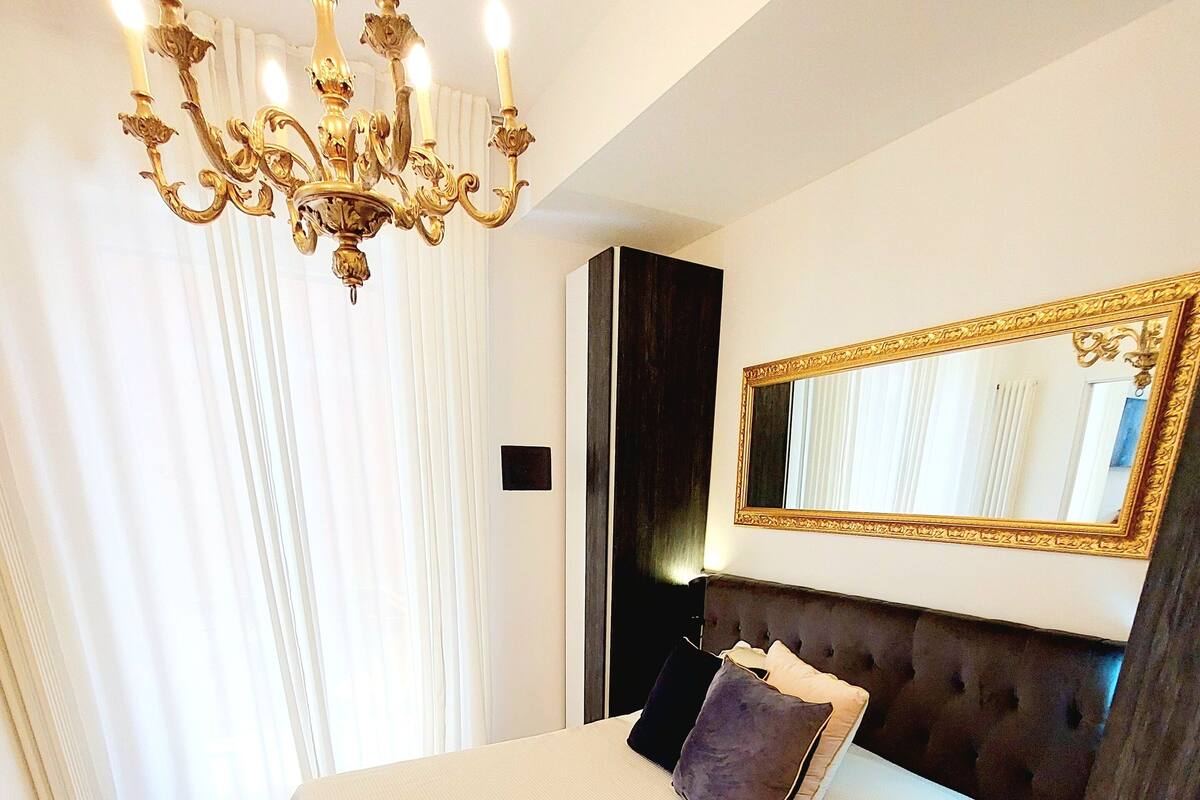 Quarto com cama grande, lustre, espelho e janela com cortinas.
