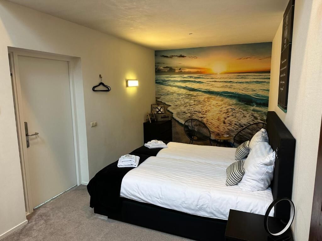 Een slaapkamer met een bed, een nachtkastje, een wandlamp, een deur en een schilderij van een strandgezicht.