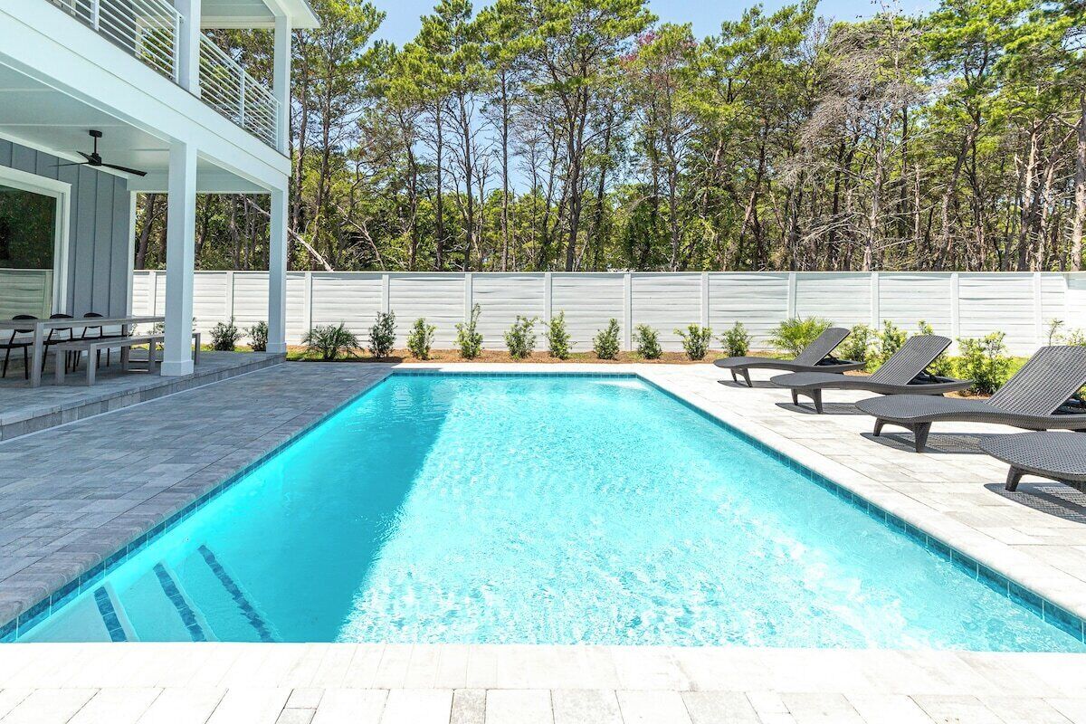 Una piscina rectangular con un deck blanco, rodeada de sillas de descanso y una cerca blanca.