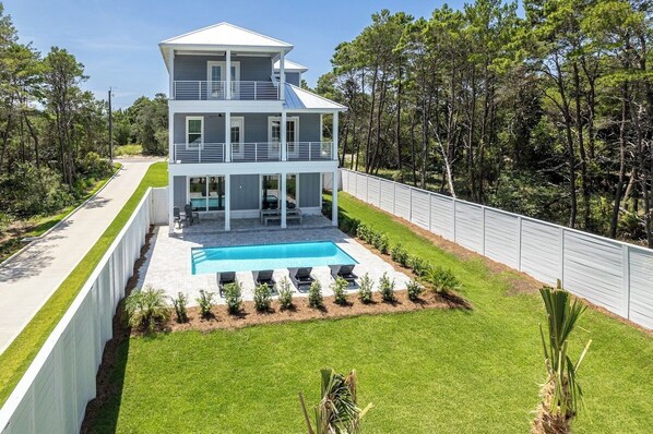 A two-story house with a pool, surrounded by a white fence and lush greenery.
