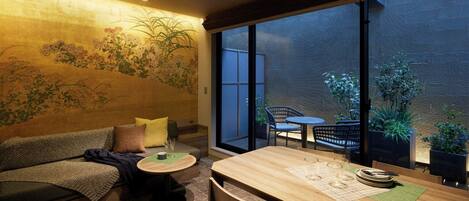 Una habitación acogedora con sofá, una pequeña mesa redonda y una mesa de comedor. Hay ventanas grandes con vistas al exterior.