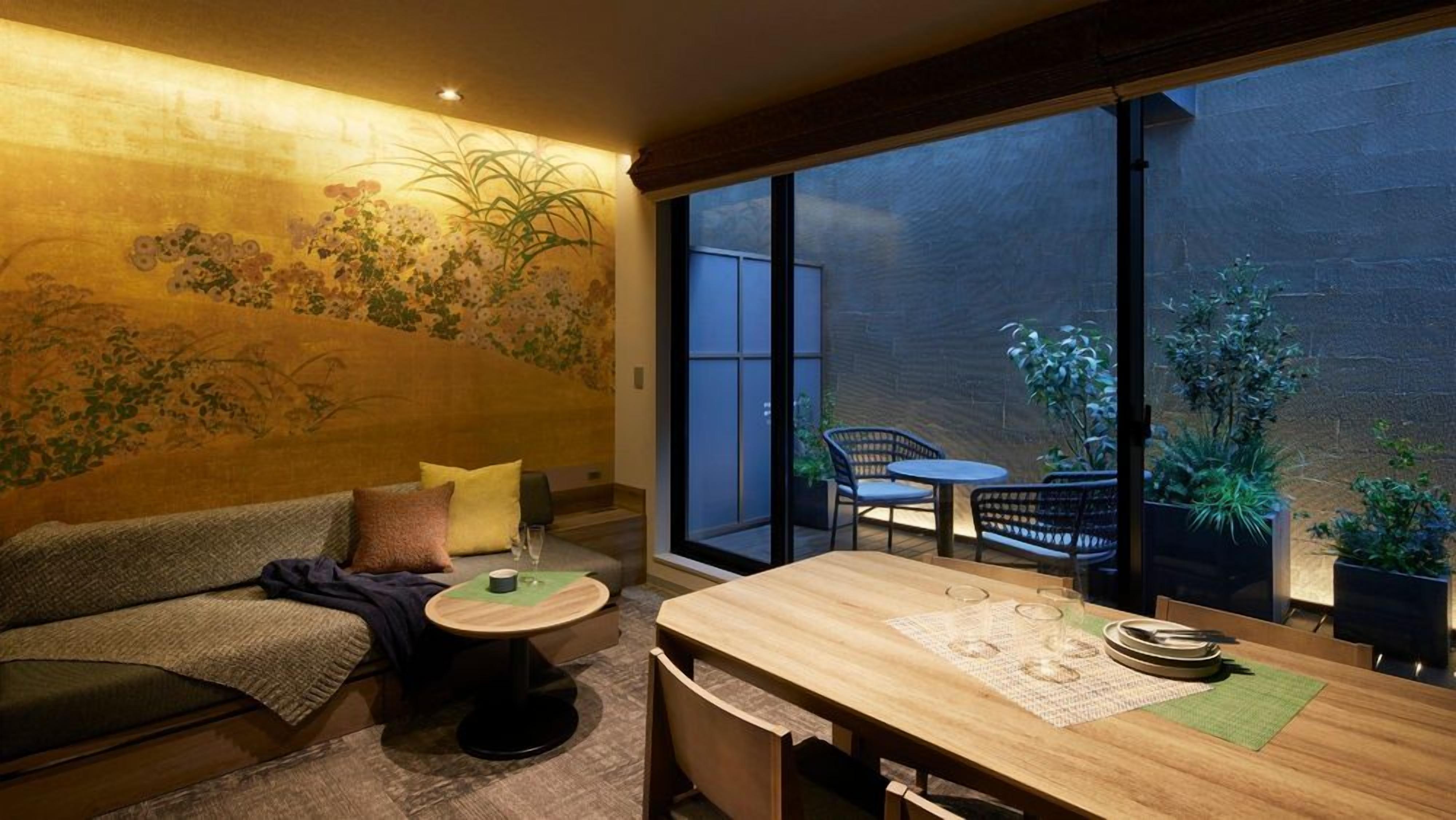 A cozy room with a sofa, a small round table, and a dining table with chairs. There is a large window providing a view of the outside.