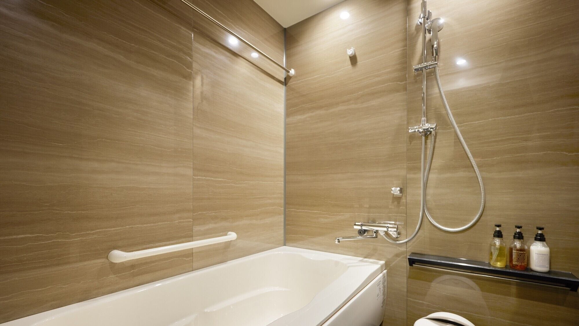 Een moderne badkamer met een badkuip, douche en toilet. Er zijn handsteunen en een plank met toiletartikelen.