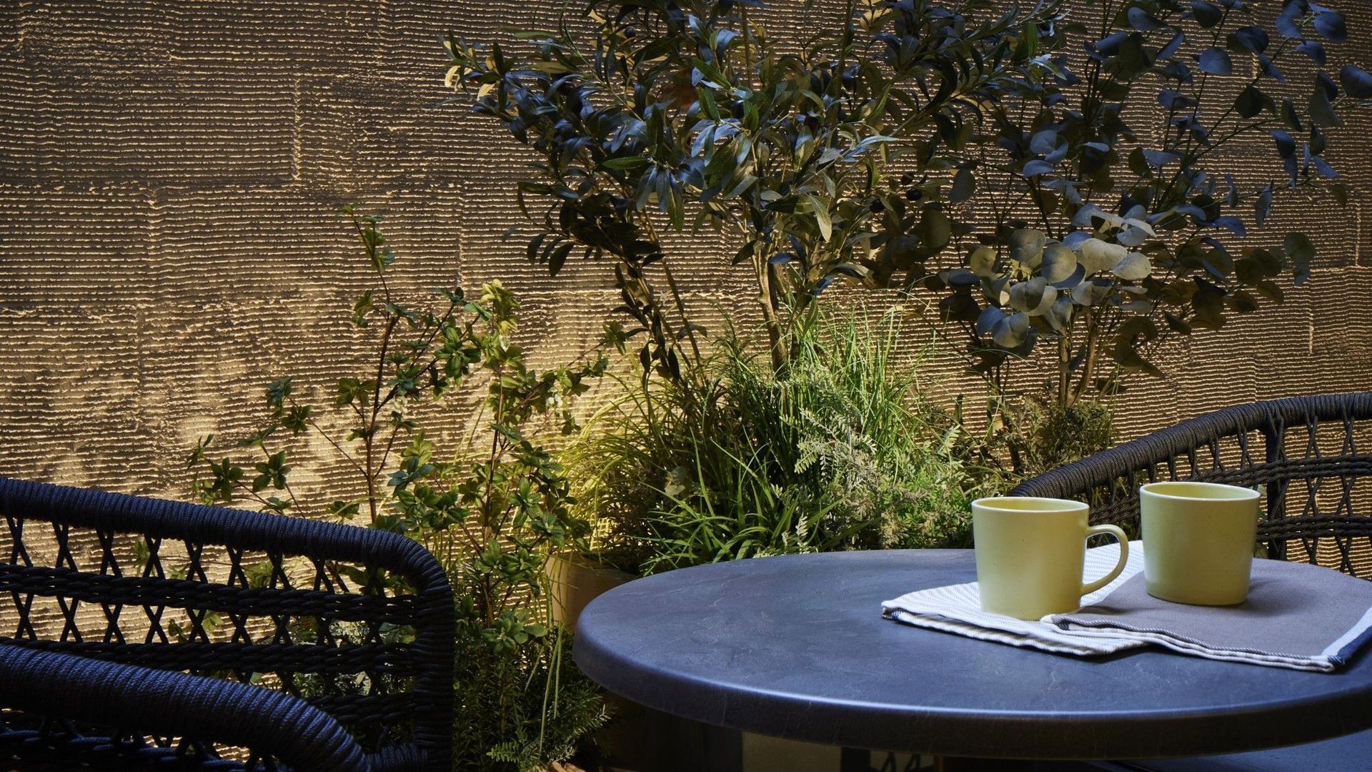 A round table with two yellow mugs on a woven mat, surrounded by wicker chairs, with a textured wall and greenery in the background.