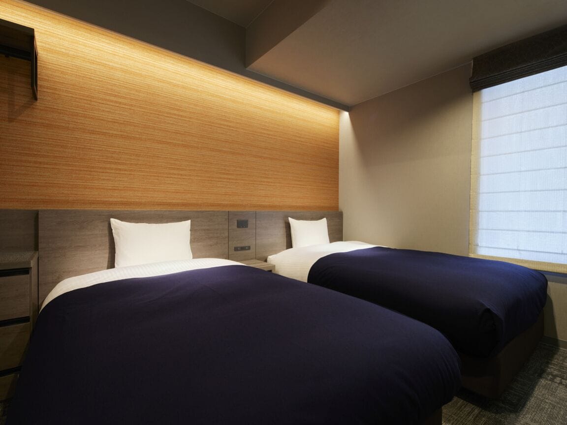 A hotel room with two beds, a wooden headboard, a window with blinds, and a nightstand.