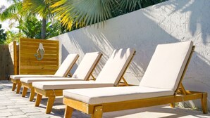 A row of wooden lounge chairs with white cushions, arranged on a paved patio with a white wall and palm trees in the background. - Another Day In Paradise - Coastal Holiday! (St. Augustine Vilano Beach)