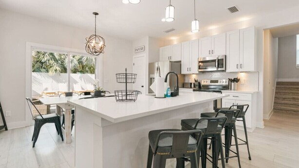 A modern kitchen with white cabinetry, a large island with bar stools, pendant lighting, and a view of the outdoors. - Another Day In Paradise - Coastal Holiday! (St. Augustine Vilano Beach)