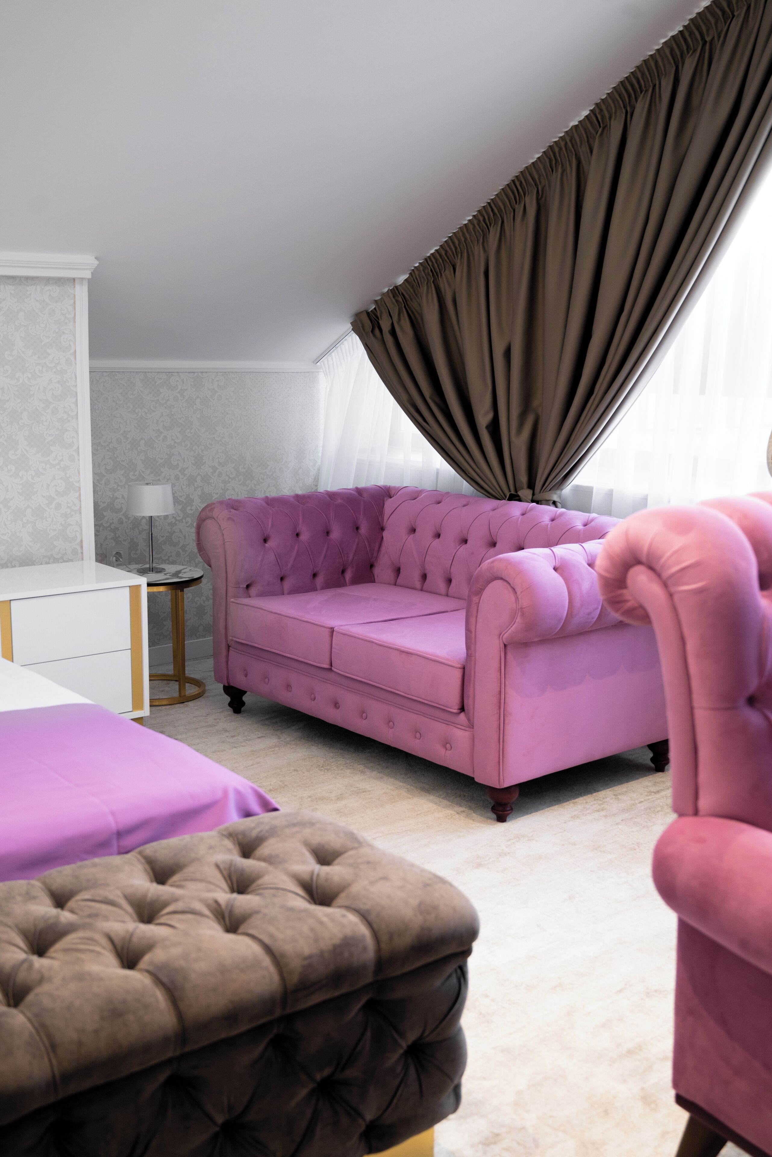 A room with a tufted pink sofa, a matching ottoman, a white bedside table, and a floor lamp.