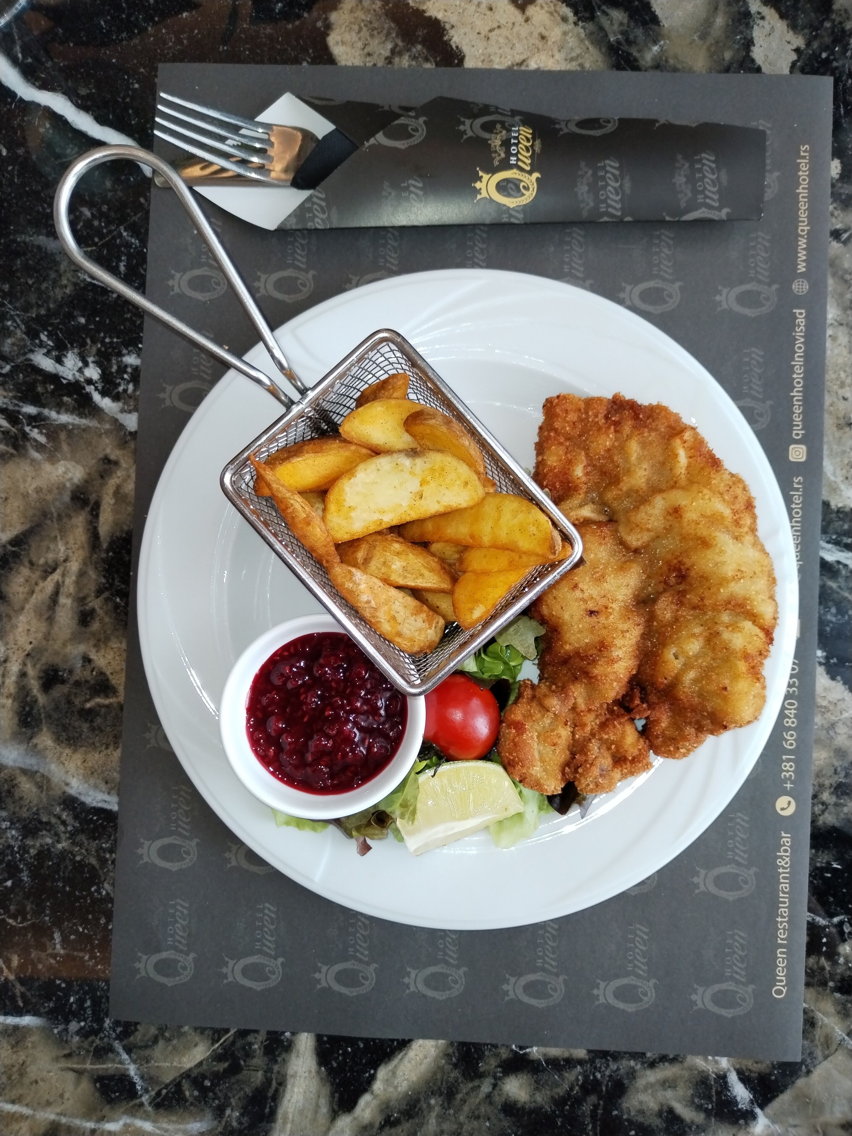 Un plat amb una escalivada empanada, patates fregides, un bol petit de salsa de cranberry, un tomàquet cherry i una llesca de llimona sobre un plat blanc.