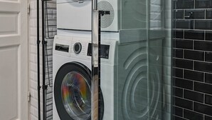 A stacked washer and dryer unit in a modern laundry room with a glass partition and black tiled backsplash. - Etoile Apartment (G1) – Luxury Two-Bedroom (Paris)