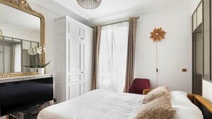 A bedroom with a bed, a dresser, a vanity, a mirror, a red chair, and a decorative wall piece. - Etoile Apartment (G1) – Luxury Two-Bedroom (Paris)