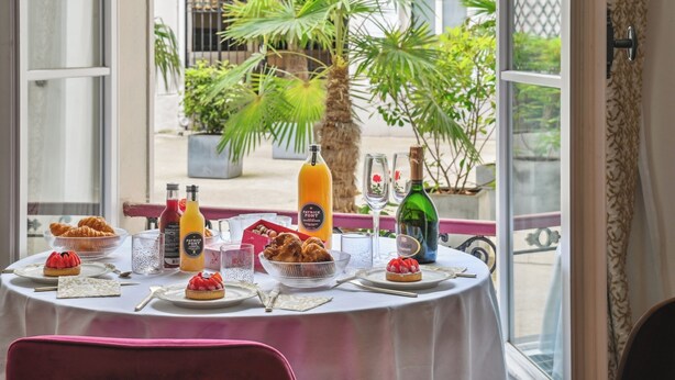 A dining table set with breakfast items, including pastries and beverages, in a room with a view of palm trees and a building outside. - Etoile Apartment (G1) – Luxury Two-Bedroom (Paris)