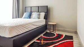 A modern bedroom with a large bed, a small stool, and a patterned rug. - 4 minutes to Two Rivers Mall! Family-sized condo with a fully functional Kitchen (Ruaka)