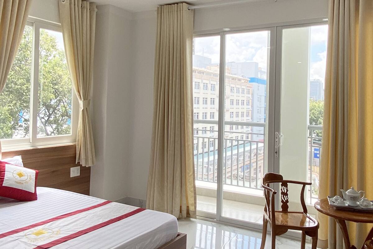 A hotel room with a bed, a chair, a table with teapots, and a balcony with a view of buildings.