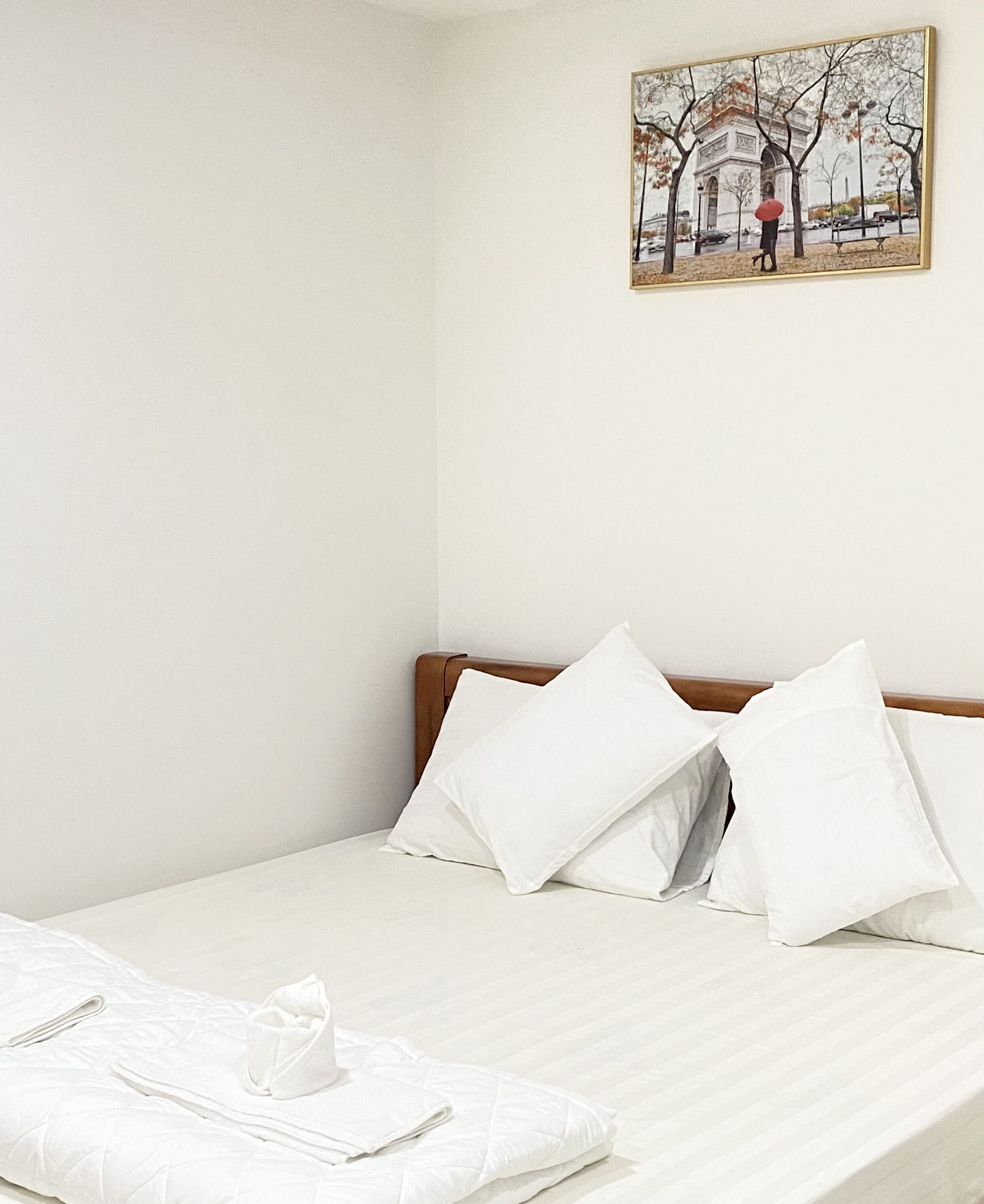 A neatly made bed with white bedding and pillows, a wooden headboard, and a framed picture of the Eiffel Tower on the wall.