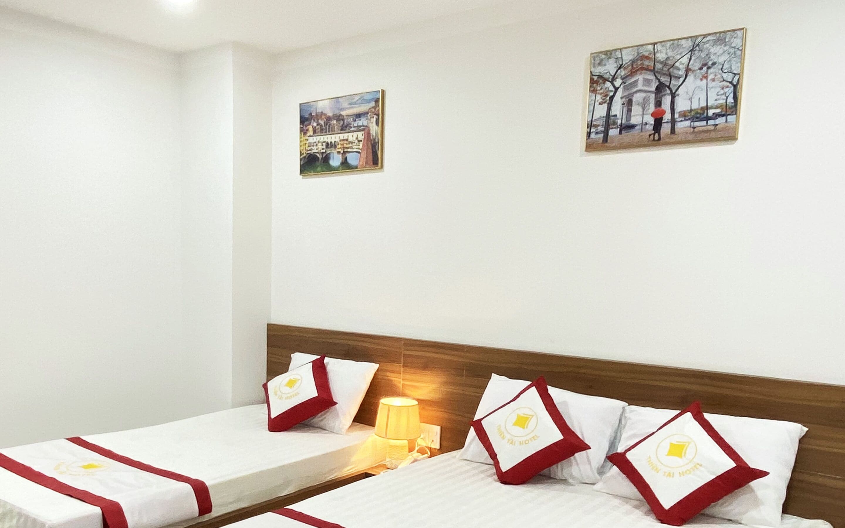 A hotel room with two single beds, each with a red and white pillow featuring the hotel logo. There is a nightstand with a lamp between the beds. The room has white walls and two paintings on the wall.