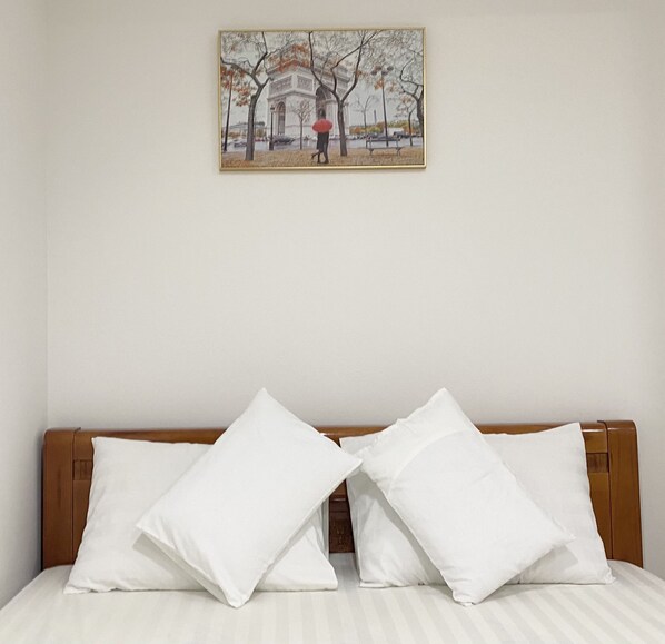 Una cama con ropa blanca y dos almohadas blancas, un cabecero de madera y un cuadro con una escena de parque en la pared.