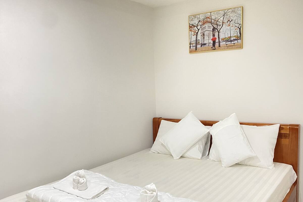 A neatly made bed with white linens and pillows, a wooden headboard, and a painting of a park scene on the wall.
