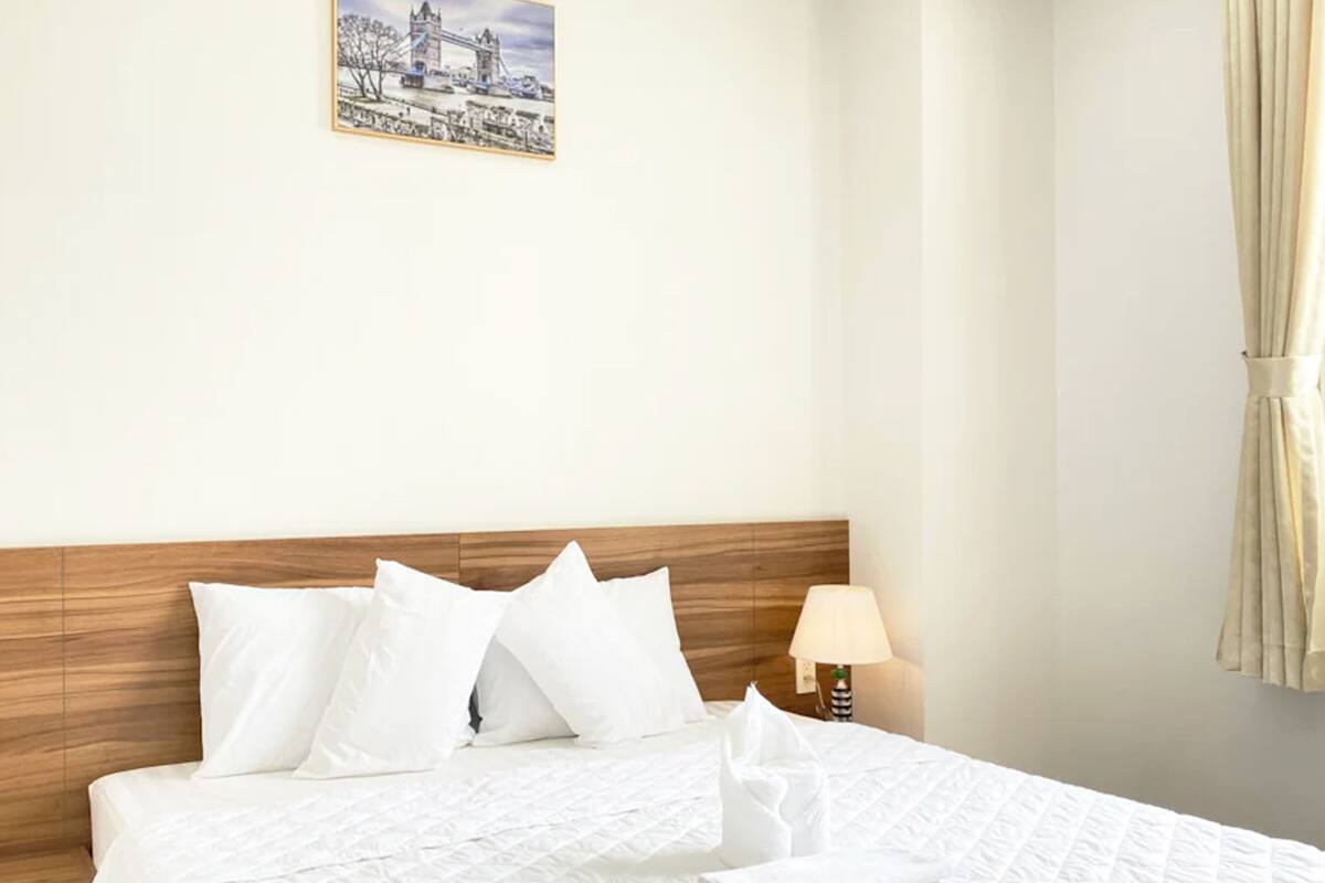 A hotel room with a wooden headboard, white bedding, a bedside lamp, and a window with curtains.