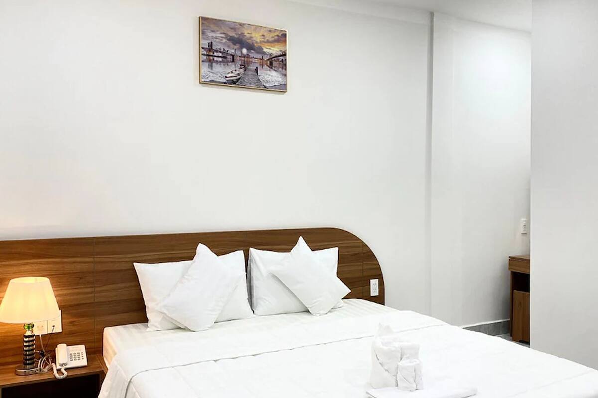 A hotel room with a wooden headboard, white bedding, a bedside lamp, a phone, and a painting on the wall.