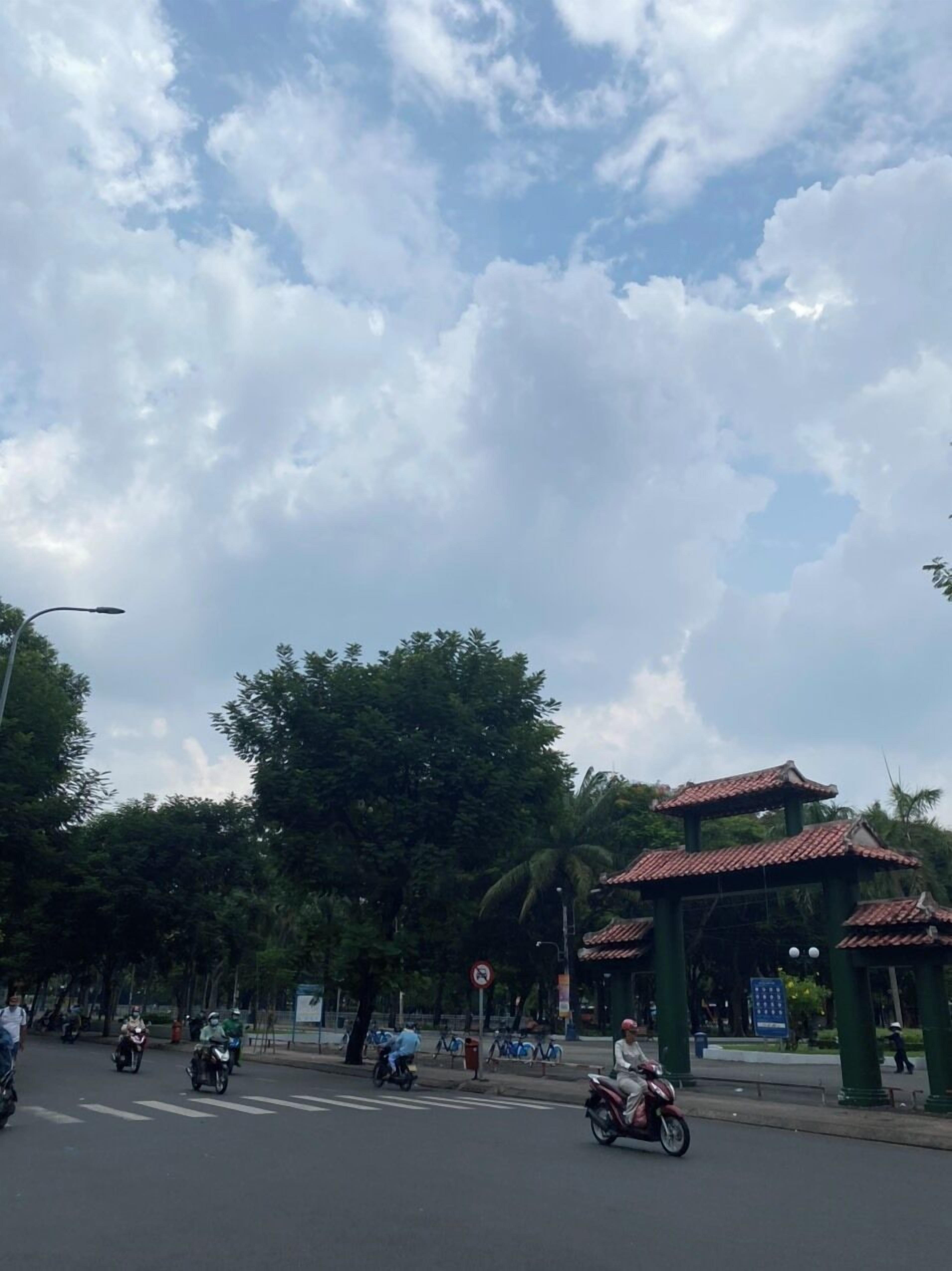 Una escena callejera con cielo despejado y nubes, una estructura con techo verde y árboles.
