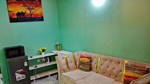 Een kamer met een groene muur, een bank met kleurrijke kussens, een kleine tafel met een bloevas en een koelkast met sticker.