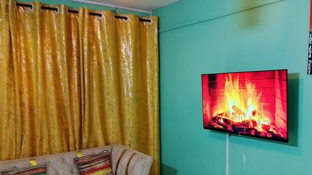 Een kamer met een groenblauwe wand, een televisie waarop een haard te zien is, en een bank met gestreepte kussens.