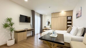 A modern hotel room with a flat-screen TV, a wooden desk, a bed with white bedding, a coffee table, and a potted plant. - CityHouse NEST LightHouse Saigon (Ho Chi Minh City)