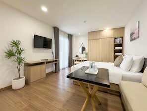A modern hotel room with a flat-screen TV, a wooden desk, a bed with white bedding, a coffee table, and a potted plant. - CityHouse NEST LightHouse Saigon (Ho Chi Minh City)