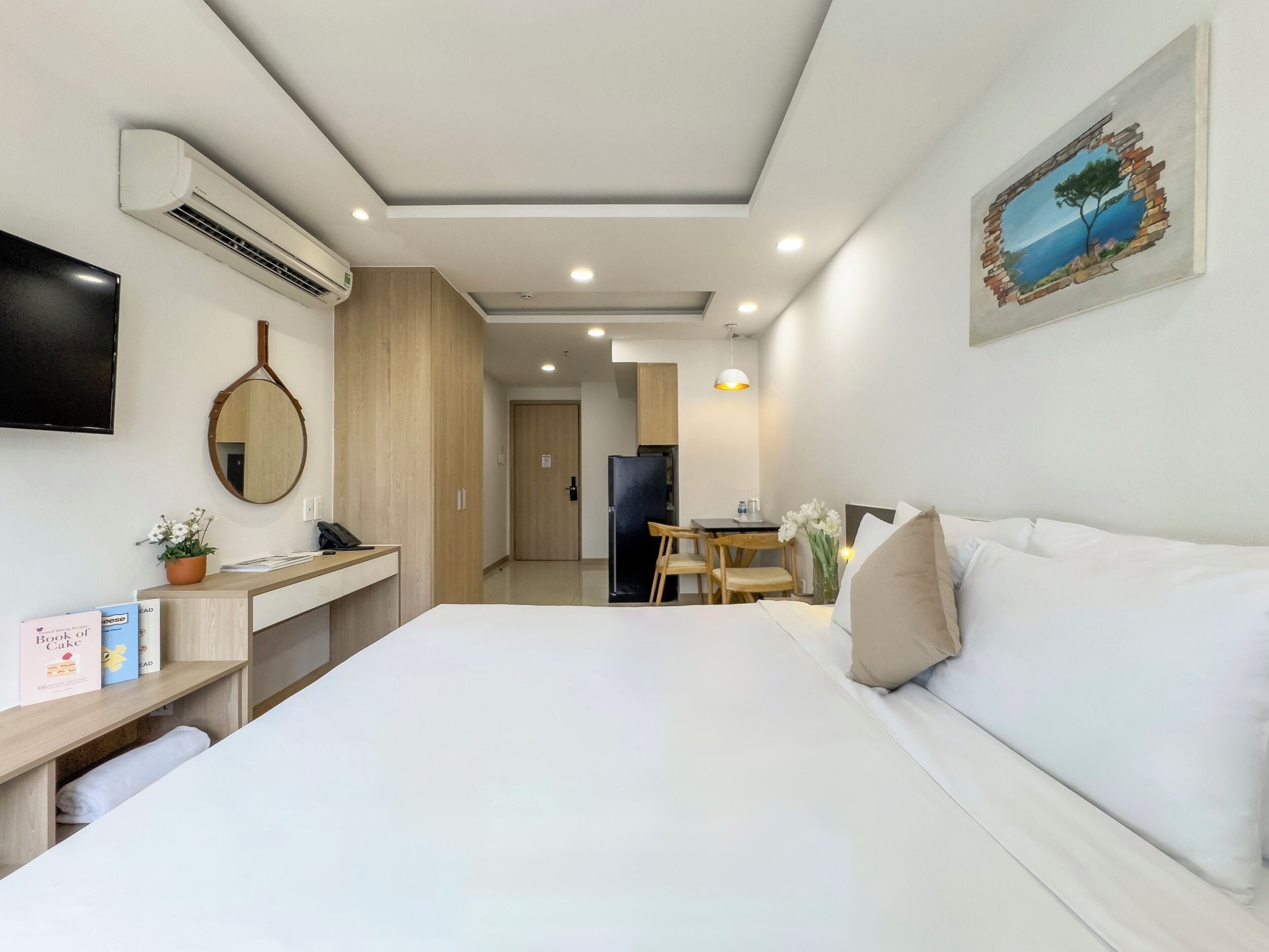 A hotel room with a bed, a desk, a mirror, an air conditioner, a TV, and a painting on the wall.