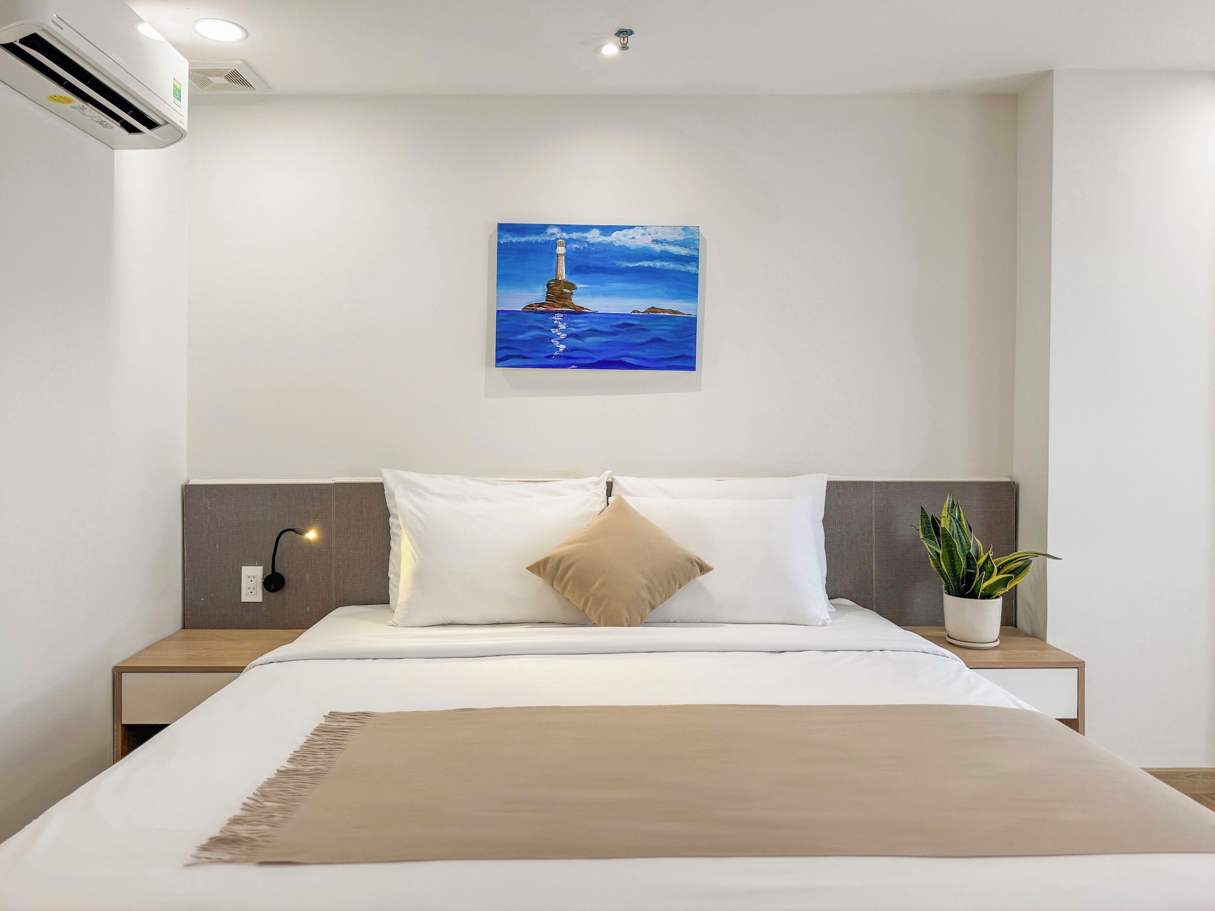 Une chambre d’hôtel avec un lit, un tableau représentant un phare, une plante en pot et un appareil de climatisation.