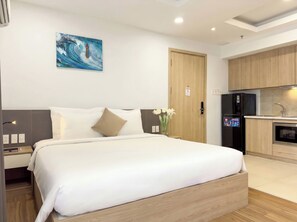 Una habitación de hotel moderna con una cama, un escritorio pequeño, un cuadro colgado en la pared, una puerta de madera, una zona de cocina con refrigerador y un lavabo.