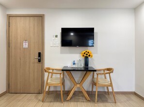 A room with a wooden door, a black table with two wooden chairs, a TV mounted on the wall, and a vase with sunflowers.