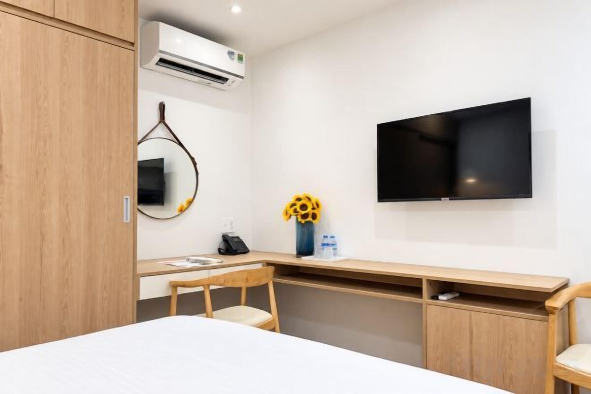 A hotel room with a bed, a desk with a chair, a flat-screen TV, an air conditioning unit, and a vase with sunflowers.