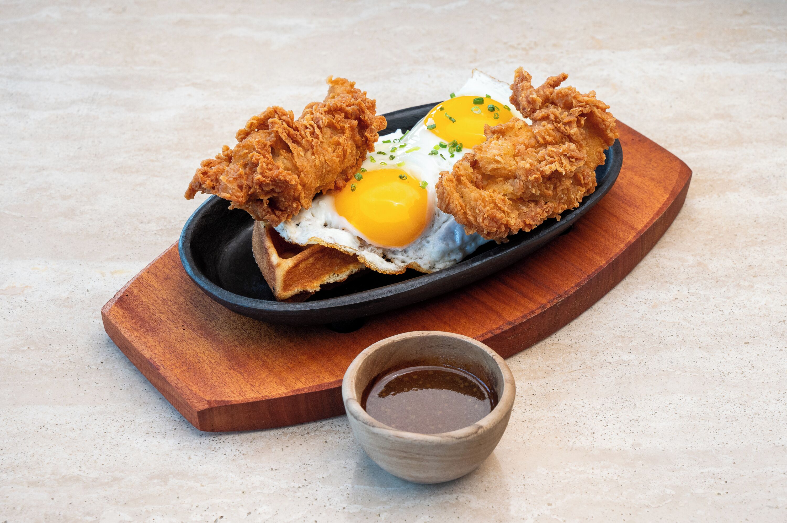 Un plato de pollo frito y huevos sobre una tabla de madera, con un recipiente pequeño de salsa al lado.