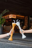 Two hands holding cocktail glasses with straws, one with a yellow base and one with a white base, in front of a building with a sign that reads "Cafe".