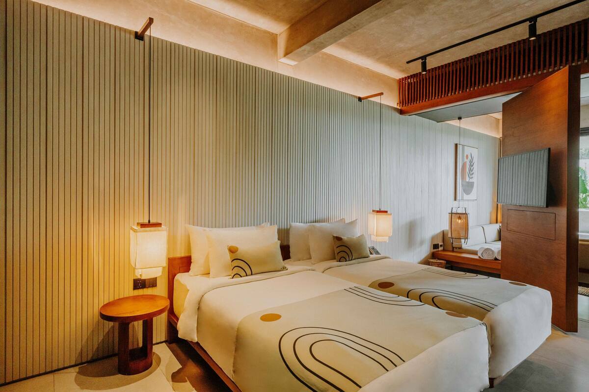 Habitación de hotel con dos camas, un banco de madera, una mesita pequeña y un televisor montado en la pared.