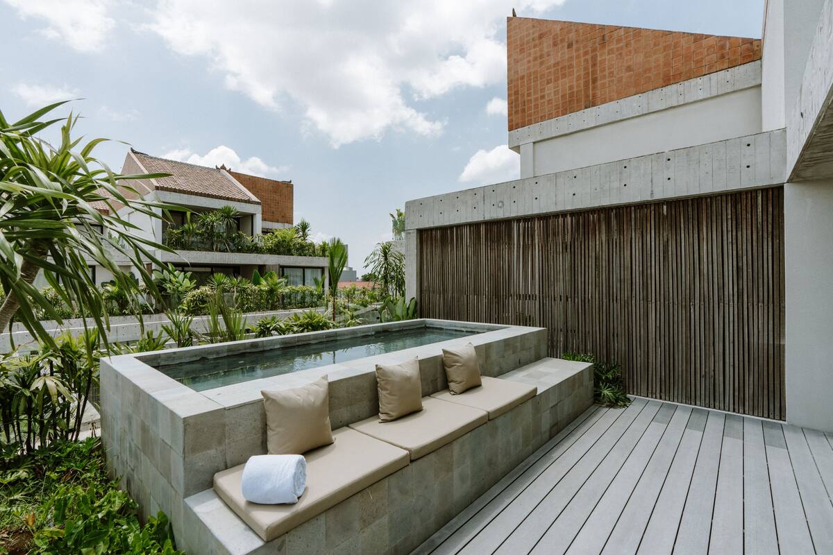 Un área de piscina exterior moderna con deck de concreto, cojines color beige y valla de madera.